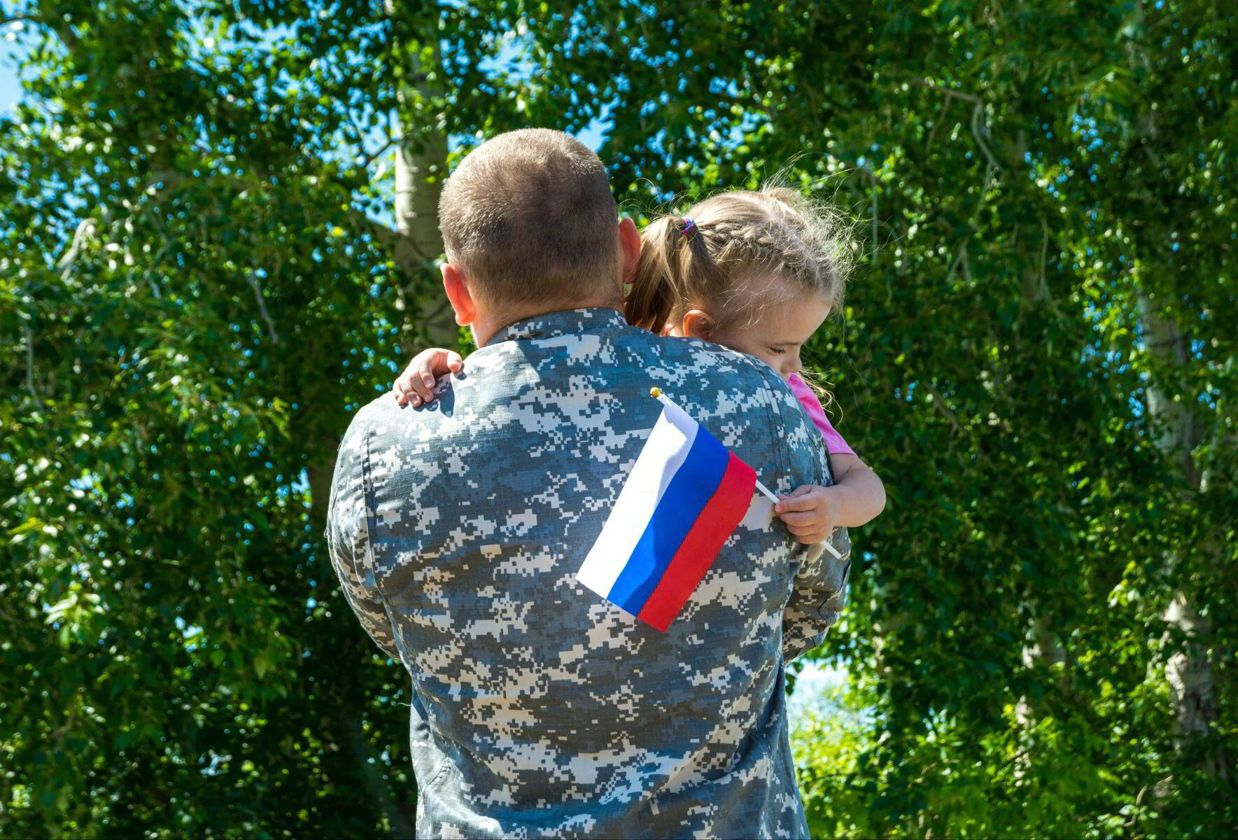 Service member in uniform hugging his daughter, who holds a small flag